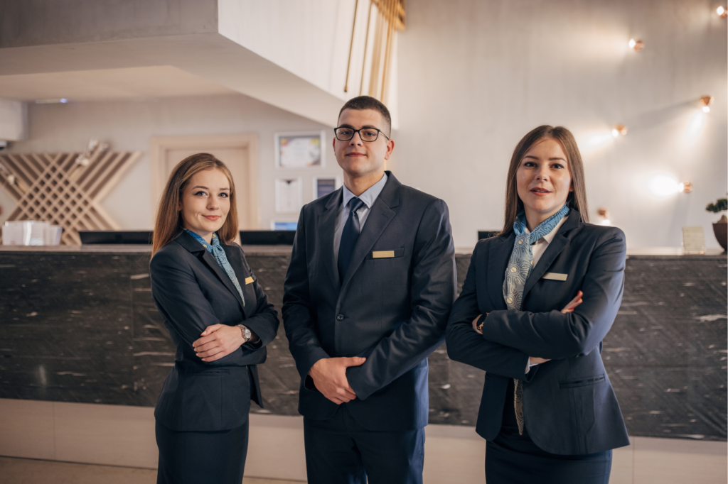 Trois professionnels de l'hospitality debout devant un comptoir d'accueil, habillés en uniformes élégants, offrant un service de qualité dans un environnement d'entreprise.