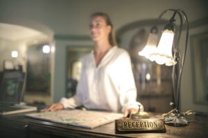 Réceptionniste accueillante incarnant l’hospitality derrière un comptoir d’hôtel avec une cloche de réception et une lampe vintage.