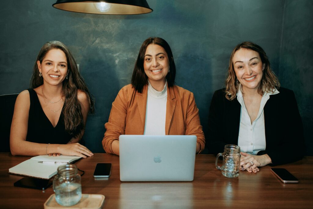 Trois femmes professionnelles souriantes discutent de hospitality management lors d'une réunion dans un environnement de travail moderne, avec un ordinateur portable et des notes sur la table.